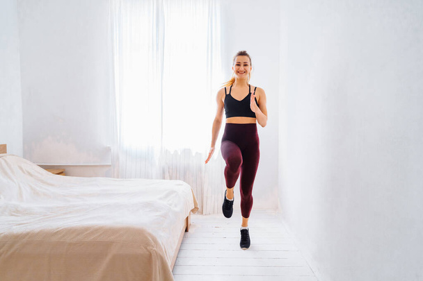 Starkes athletisches Fit-Mädchen in Top und Leggings joggt und hüpft energisch lächelnd in ihrem geräumigen und hellen Schlafzimmer mit minimalistischem weißen Interieur. - Foto, Bild