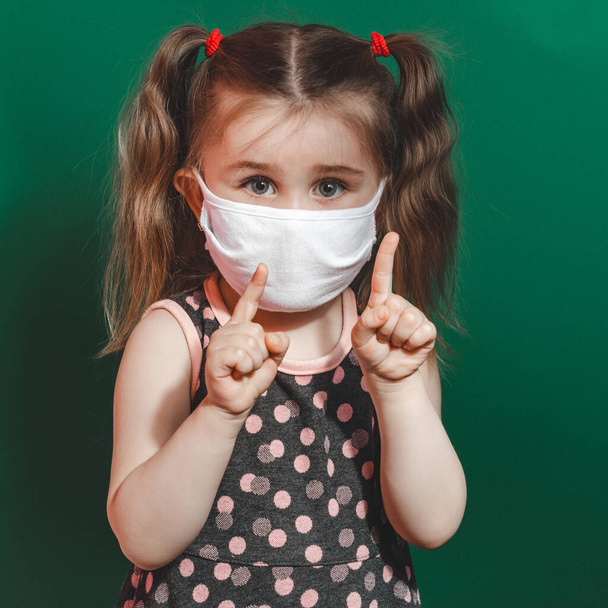 Εννοιολογική φωτογραφία του μικρού κοριτσιού με ιατρική μάσκα που δείχνει το δάχτυλο στο πράσινο φόντο κοντά στο 2020 - Φωτογραφία, εικόνα