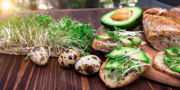 アボカドとマイクログリーンのサンドイッチ木の背景にウズラの卵。健康的な食事。選択肢だ。朝食を作る過程で. - 写真・画像