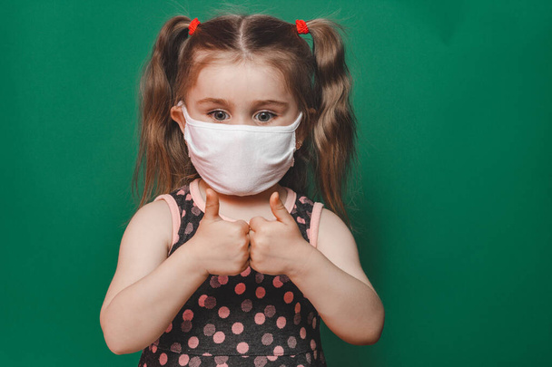 Kis kaukázusi lány orvosi maszkban és pöttyös ruhában, hüvelykujj jelet mutat zöld alapon a karantén és a koronavírus világjárvány idején 2020-ban - Fotó, kép