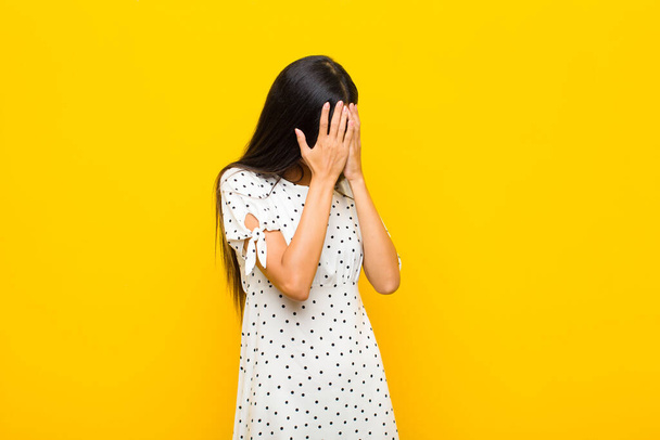 young pretty latin woman covering eyes with hands with a sad, frustrated look of despair, crying, side view against flat wall - Фото, изображение