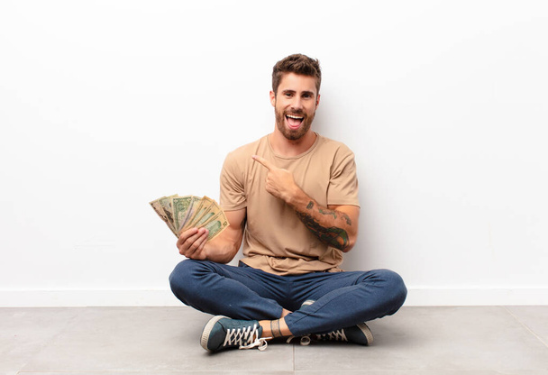 looking excited and surprised pointing to the side and upwards to copy space holding dollar banknotes - Fotoğraf, Görsel