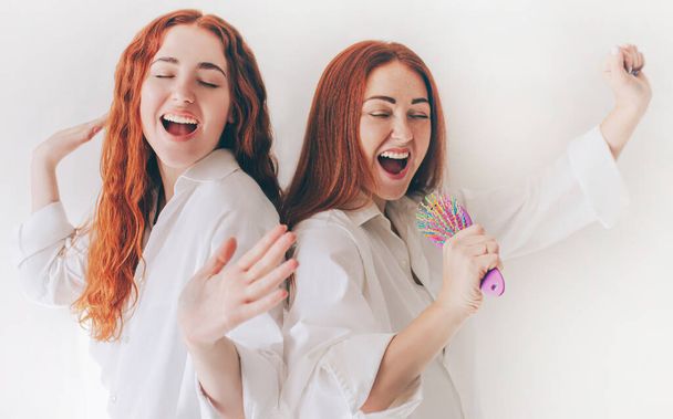 Dos mujeres jóvenes cantan en micrófonos improvisados - cepillos para el cabello. Dos hermanas de pelo rojo están aisladas sobre un fondo blanco con amplias camisas de gran tamaño
. - Foto, imagen