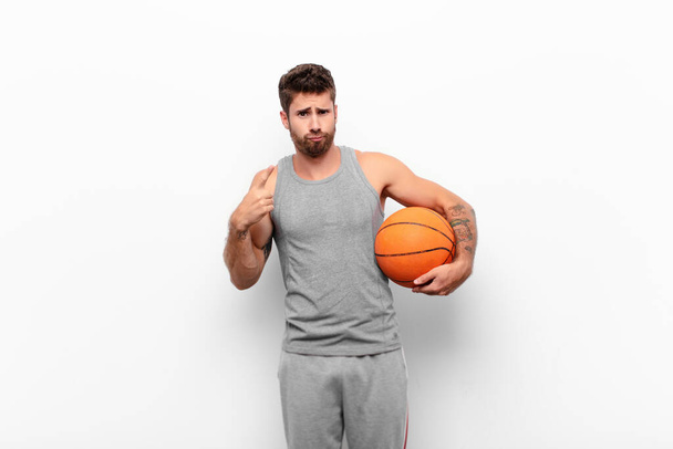 young handsome man with a bad attitude looking proud and aggressive, pointing upwards or making fun sign with hands holding a basketball ball. - Фото, изображение