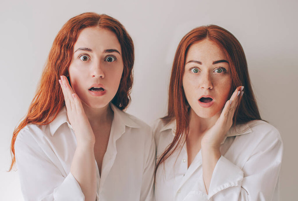 Две рыжеволосые сестры стоят изолированно на белом фоне в просторных огромных рубашках. Они очень удивлены. Они были шокированы происходящим.
. - Фото, изображение