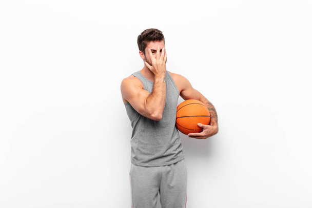 young handsome man feeling scared or embarrassed, peeking or spying with eyes half-covered with hands holding a basketball ball. - Foto, Imagem