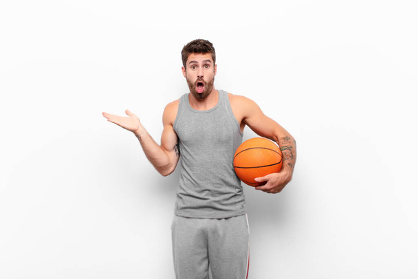 young handsome man looking surprised and shocked, with jaw dropped holding an object with an open hand on the side holding a basketball ball. - Fotoğraf, Görsel