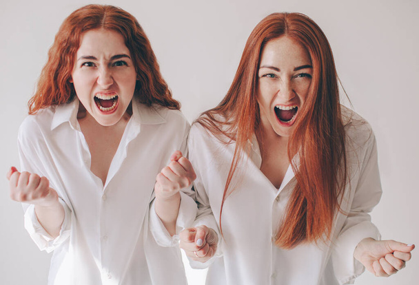 Δύο νεαρές γυναίκες θύμωσαν και φώναξαν στην κάμερα αισθάνονται οργή, επιθετικότητα, θυμό. Δύο κοκκινομάλλες αδελφές στέκονται απομονωμένες σε ένα λευκό φόντο σε ευρύχωρα υπερμεγέθη πουκάμισα - Φωτογραφία, εικόνα