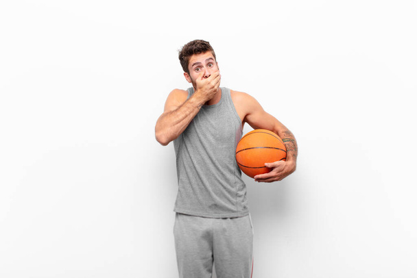 young handsome man covering mouth with hands with a shocked, surprised expression, keeping a secret or saying oops holding a basketball ball. - Fotografie, Obrázek