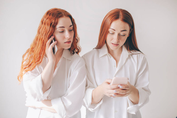 Two red-haired sisters stand isolated on a white background in spacious oversized shirts. Two young women are addicted to social networks spend all their time in a smartphone - Photo, Image