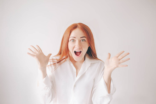 Een jonge, roodharige vrouw geïsoleerd op een witte achtergrond in een wit T-shirt heeft een schok van verrassing, verrukking en vreugde. Ze deed haar mond open en schreeuwde naar de camera.. - Foto, afbeelding