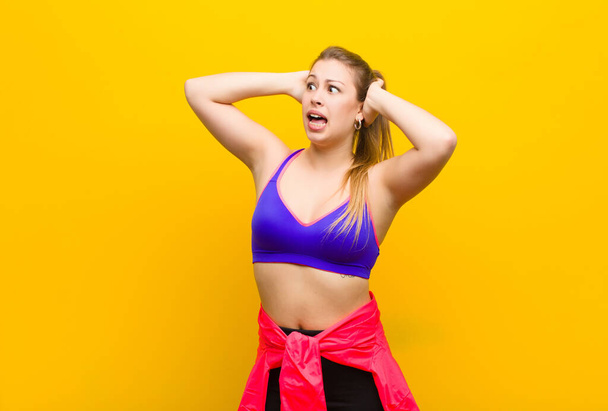 young blonde woman with open mouth, looking horrified and shocked because of a terrible mistake, raising hands to head. sport concept - Foto, imagen