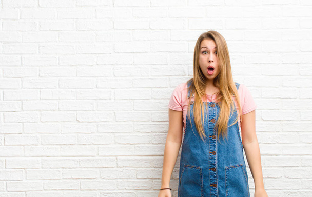 young blonde woman looking very shocked or surprised, staring with open mouth saying wow against brick wall - Foto, immagini