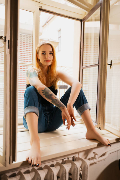 Blonde woman sits on windowsill wearing black t-shirt and jeans and looks at you - Fotoğraf, Görsel