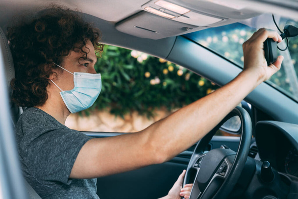 Tıbbi maskeli ve gri tişörtlü genç kıvırcık adam araba kullanıyor. Koruyucu maskeli bir adam bir arabanın direksiyonunda oturuyor. Koronavirüs salgınından korunma, kovid-19. Yeni gerçeklik - Fotoğraf, Görsel