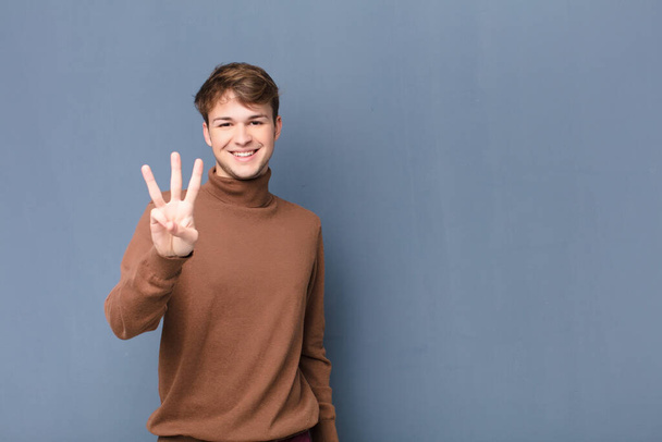 young blonde man smiling and looking friendly, showing number three or third with hand forward, counting down isolated against flat wall - Valokuva, kuva