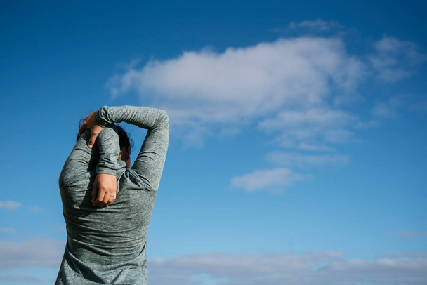 mladá žena zezadu protahuje ramena po cvičení, s oblohou v pozadí - Fotografie, Obrázek