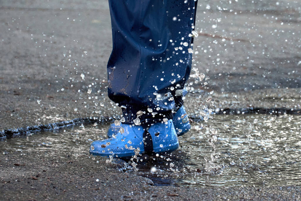 Gelukkig kind springen in plas in waterdichte jas. Een jongen heeft plezier in het regenweer in een heldere regenjas. Benen van kind met rubberen laarzen. - Foto, afbeelding
