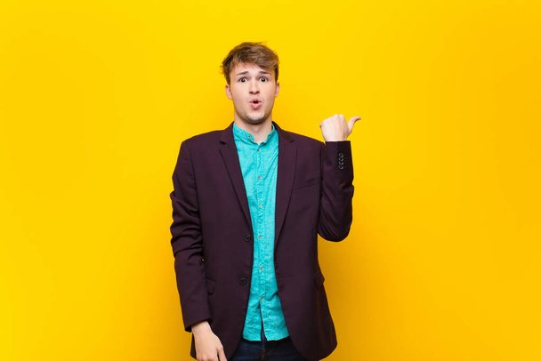 young blonde man looking astonished in disbelief, pointing at object on the side and saying wow, unbelievable isolated against flat wall - Foto, imagen