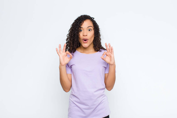 young black woman feeling shocked, amazed and surprised, showing approval making okay sign with both hands against white wall - Фото, изображение