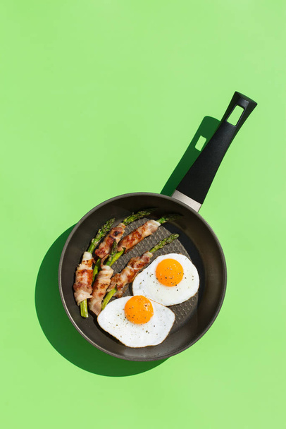 Balanced diet nutrition keto food concept. Healthy ketogenic low carb fried eggs, bacon, asparagus in a cooking pan. Trendy sunny, vivid, harsh and hard light and shadows on a green mint background - Фото, зображення