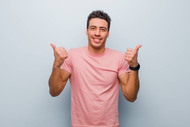 young arabian man smiling joyfully and looking happy, feeling carefree and positive with both thumbs up against gray wall - Фото, зображення