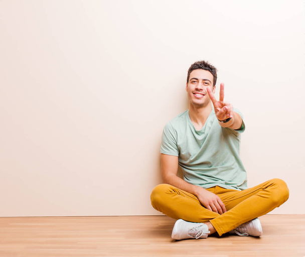 young arabian man smiling and looking happy, carefree and positive, gesturing victory or peace with one hand sitting on the floor - Фото, зображення