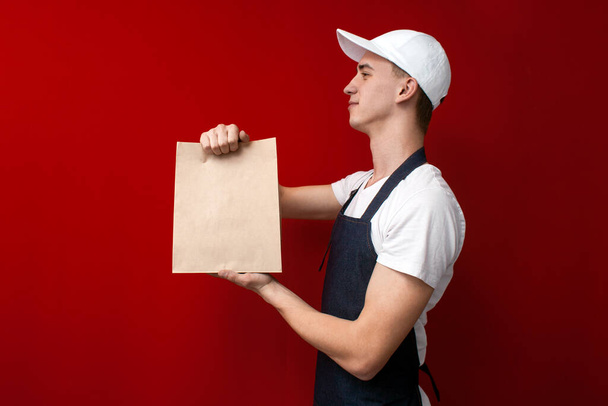 joven mensajero alegre da paquetes con una orden y sonríe, repartidor sostiene paquetes sobre un fondo rojo
 - Foto, imagen