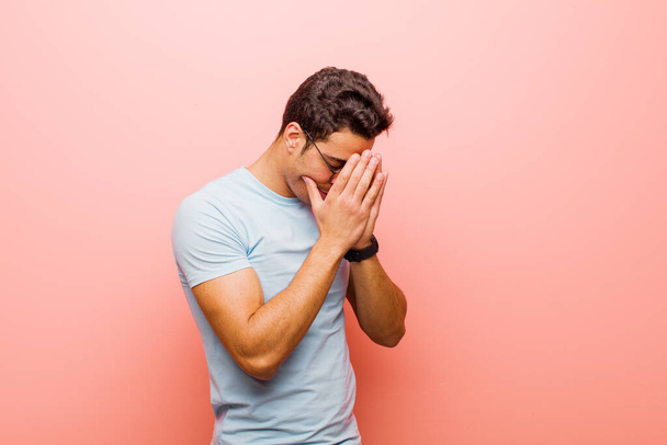 young arabian man covering eyes with hands with a sad, frustrated look of despair, crying, side view against pink wall - Фото, изображение