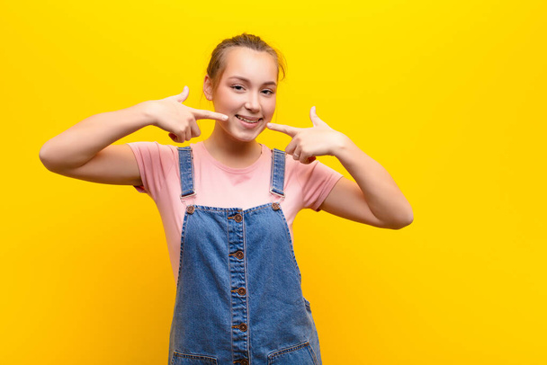 young blonde pretty girl smiling confidently pointing to own broad smile, positive, relaxed, satisfied attitude against yellow wall - Zdjęcie, obraz
