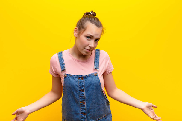 young blonde pretty girl feeling clueless and confused, having no idea, absolutely puzzled with a dumb or foolish look against yellow wall - Fotoğraf, Görsel