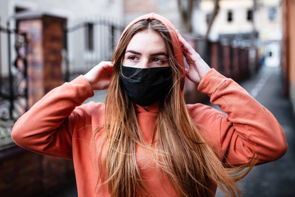 Een jong meisje met rood haar in een zwart medisch masker loopt tijdens een coronavirusepidemie. Pandemie Coronavirus COVID-19 Een jong meisje op straat met een gezichtsmasker. Meisje met een capuchon. Roodharig meisje. - Foto, afbeelding