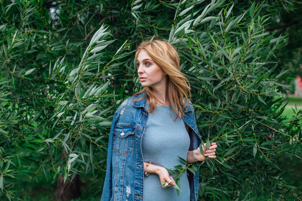 Portrait of a young beautiful woman with make up and curly blond hair dressed in jeans jacket, standing next to the tree with green leaves in a windy weather. Copy space. - Foto, imagen