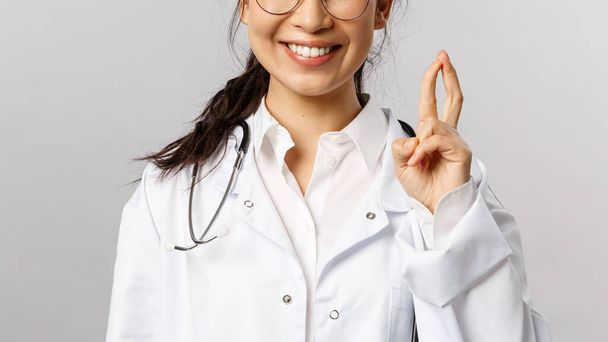 Detailní záběr portrét nadšené, usmívající se atraktivní asijské ženy lékař, terapeut check-up dítě v nemocnici, ukazuje málo triků k povzbuzení pacienta, stojící pozitivní šedé pozadí - Fotografie, Obrázek