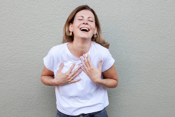 Straatfoto van schattige blanke vrouw in witte t-shert die plezier hebben hardop lachen van vreugde en grappige grap gebaren met palm lachend en lachend zorgeloos op camera - Foto, afbeelding