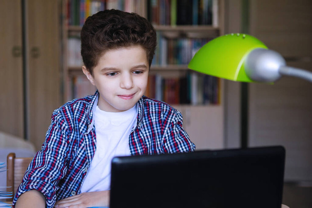 A tinédzser fiú az asztalnál laptoppal örömmel néz a monitorra. A koronavírus karantén alá helyezte az otthoni oktatást. Szelektív fókusz. Homályos háttér. - Fotó, kép