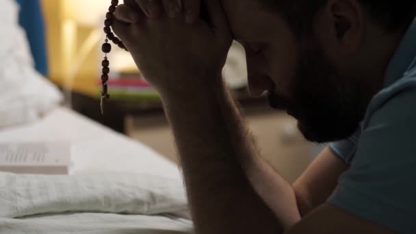 De mens is aan het bidden. Baard man zit op de vloer in de slaapkamer door bed, houdt rozenkrans met kruisbeeld in zijn handen en zegt gebed. Close-up zicht - Video