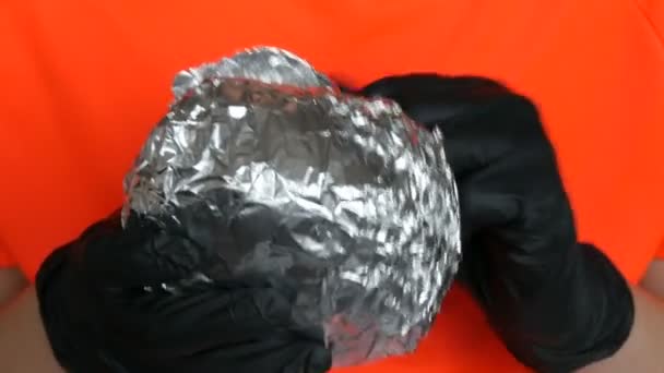 Een tiener ontvouwt grote folie hamburger. Handen in speciale zwarte handschoenen houden junkfood vast, fastfood. Gezicht en mond close-up uitzicht. - Video