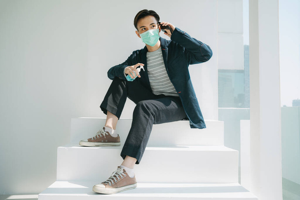 Fashion portrait - Young asian thai man wearing doctor mask using smartphone and cleaning it with alcohol gel. - Photo, Image
