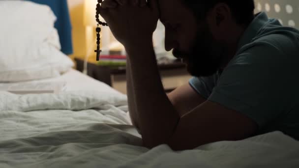 Człowiek się modli. Brodaty mężczyzna siedzi na piętrze w sypialni przez łóżko, trzyma różaniec z krucyfiksem w rękach i odmawia modlitwę. Średni widok strzału - Materiał filmowy, wideo