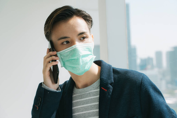 Cuarentena en el hogar concepto - Joven tailandés asiático con máscara de médico usando teléfono inteligente para hacer negocios en hone
. - Foto, imagen