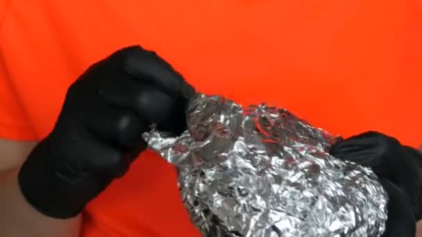 Een tiener ontvouwt grote folie hamburger. Handen in speciale zwarte handschoenen houden junkfood vast, fastfood. Gezicht en mond close-up uitzicht. - Video