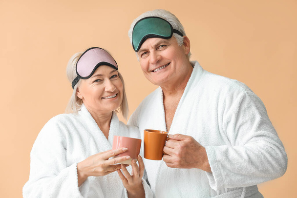 volwassen paar in badjassen en met slaap maskers drinken thee op kleur achtergrond - Foto, afbeelding