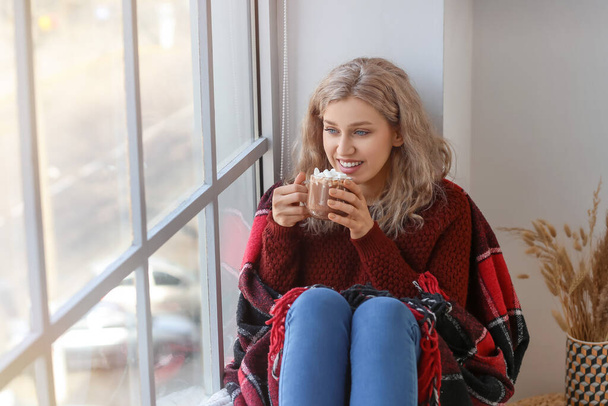 Όμορφη νεαρή γυναίκα πίνοντας ζεστή σοκολάτα κοντά στο παράθυρο στο σπίτι - Φωτογραφία, εικόνα