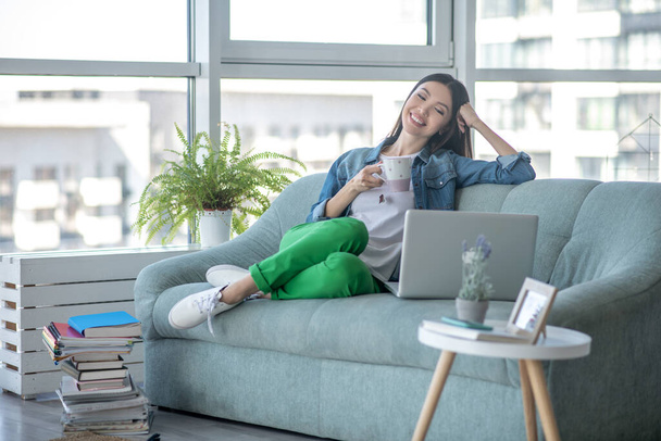 Young woman in a jeans jacket sitting on the sofa and having tea - Foto, Imagem