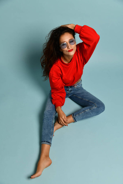 Curly brunette woman in smoked blue glasses, red sweater and blue jeans is sitting with her eyes closed feeling bliss - Foto, imagen