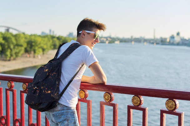 Outdoor portrait of smiling young male teenager with fashionable haircut standing on bridge over river on sunny summer day, copy space - Valokuva, kuva