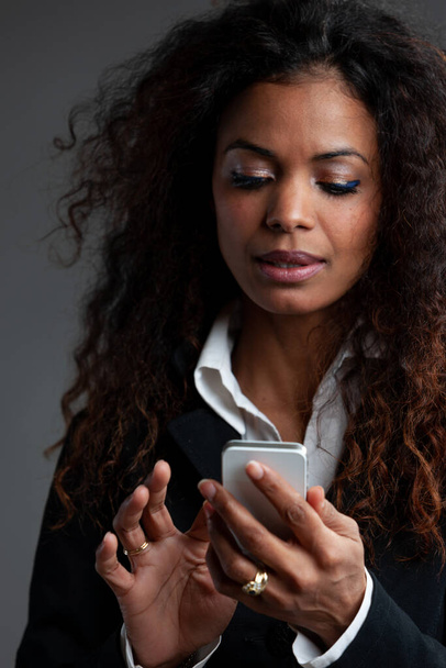 Красива жінка з довгим кучерявим темним волоссям, що стоїть на своєму портативному мобільному телефоні з продуманим виразом крупним планом на сірому
 - Фото, зображення
