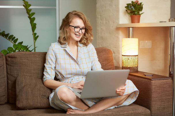 A woman in a dress sitting on the couch with legs crossed works remotely on a laptop in her apartment. A girl with braces watching a webinar. A female student in glasses listening to an online lecture - Fotografie, Obrázek