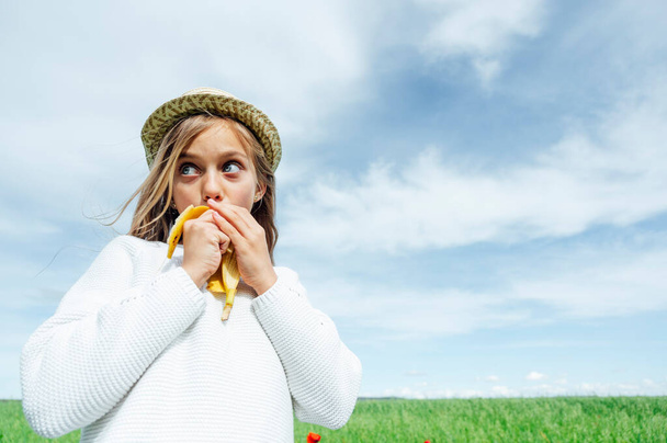 Little blond girl eating a banana in the field - Zdjęcie, obraz